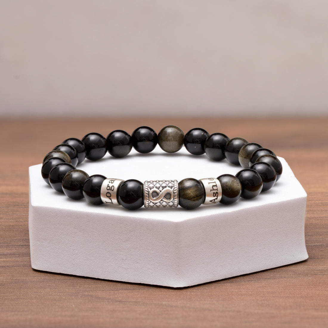 Engraved silver charms with names on a black obsidian bracelet, highlighted by a silver infinity charm, symbolizing lasting connections